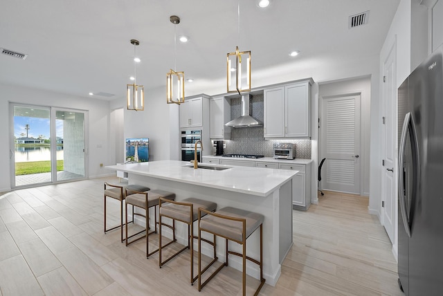 kitchen featuring pendant lighting, wall chimney range hood, a kitchen island with sink, backsplash, and stainless steel appliances