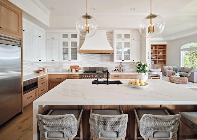 kitchen with premium range hood, sink, white cabinets, hanging light fixtures, and built in appliances