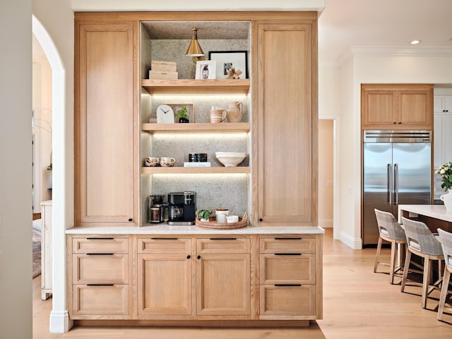 bar with pendant lighting, crown molding, built in refrigerator, light brown cabinetry, and light wood-type flooring