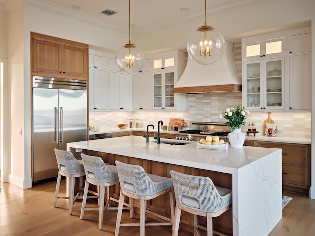 kitchen featuring premium range hood, white cabinetry, a kitchen island with sink, stainless steel built in fridge, and light wood-type flooring