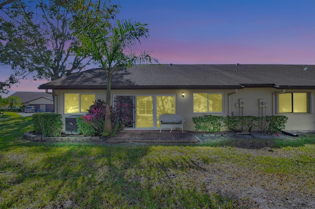 back of property at dusk featuring roof with shingles, central air condition unit, a lawn, and stucco siding