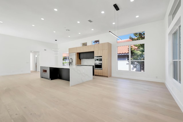 kitchen with a kitchen island with sink, light stone counters, light hardwood / wood-style floors, light brown cabinetry, and decorative light fixtures