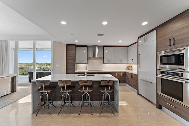 kitchen with sink, built in appliances, a center island with sink, a kitchen breakfast bar, and wall chimney range hood