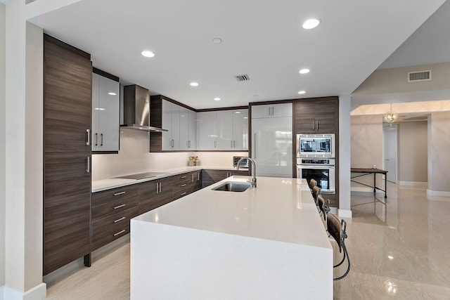 kitchen with dark brown cabinetry, a breakfast bar, sink, stainless steel appliances, and wall chimney range hood