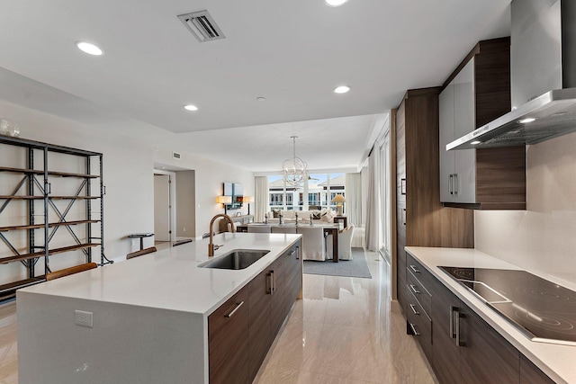 kitchen featuring wall chimney range hood, sink, a kitchen island with sink, black electric cooktop, and decorative light fixtures