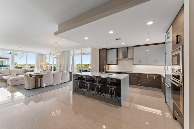 kitchen with appliances with stainless steel finishes, a kitchen island with sink, an inviting chandelier, dark brown cabinetry, and wall chimney exhaust hood