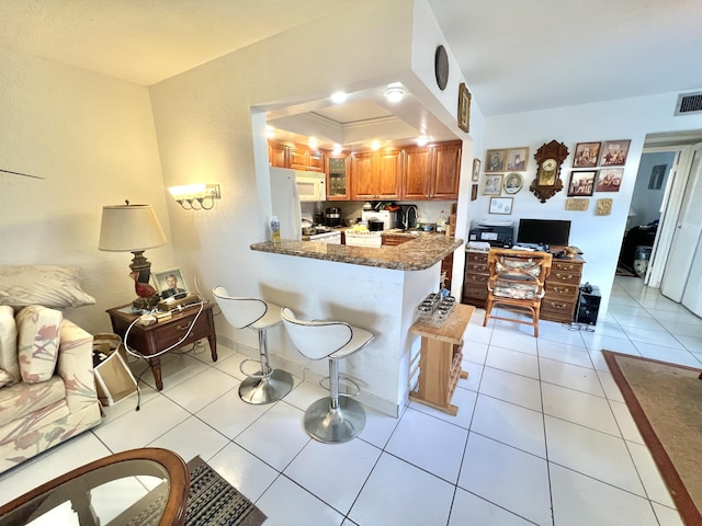 kitchen with light tile patterned floors, white appliances, a breakfast bar, kitchen peninsula, and dark stone counters