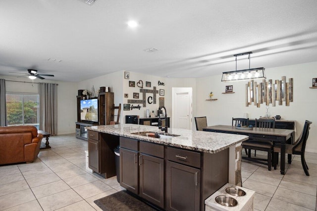 kitchen with sink, hanging light fixtures, light stone counters, dark brown cabinets, and a center island with sink