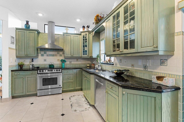 kitchen featuring sink, light tile patterned floors, appliances with stainless steel finishes, dark stone counters, and wall chimney range hood