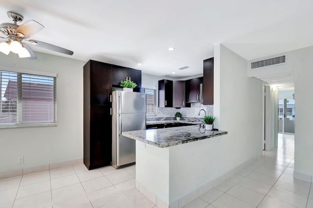 kitchen featuring stainless steel fridge, dark stone countertops, dark brown cabinetry, tasteful backsplash, and kitchen peninsula