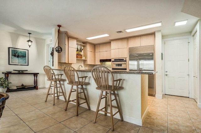 kitchen with light brown cabinetry, dark stone countertops, a kitchen bar, kitchen peninsula, and stainless steel appliances