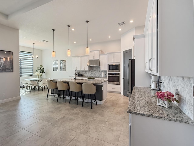 kitchen featuring light stone counters, decorative light fixtures, an island with sink, stainless steel appliances, and white cabinets