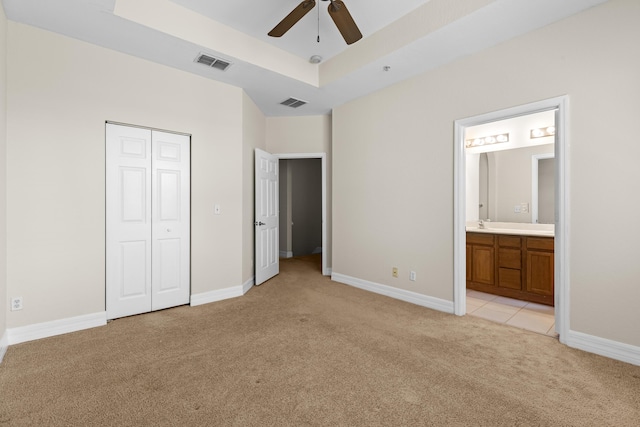 unfurnished bedroom with ensuite bath, ceiling fan, a tray ceiling, light colored carpet, and a closet