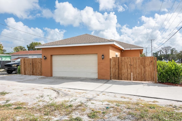 view of side of home with fence, an attached garage, a shingled roof, stucco siding, and concrete driveway