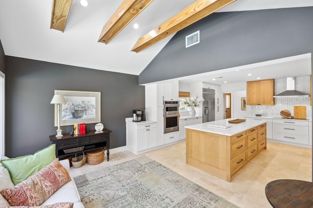 kitchen with a kitchen island, appliances with stainless steel finishes, beamed ceiling, white cabinetry, and wall chimney range hood