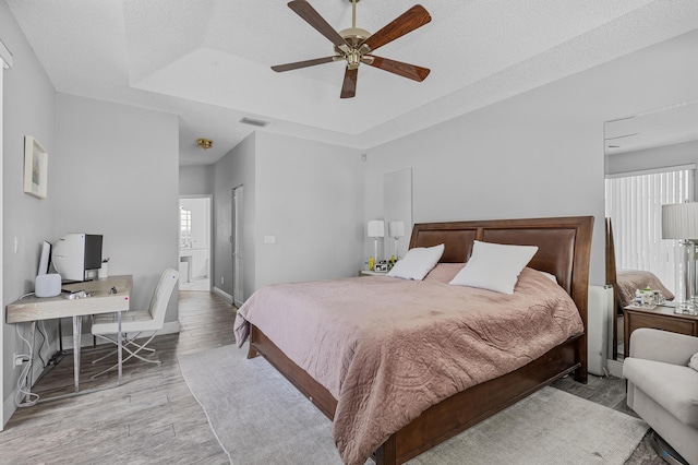 bedroom with ensuite bathroom, a tray ceiling, ceiling fan, light hardwood / wood-style floors, and a textured ceiling