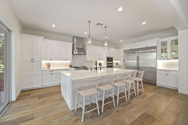 kitchen featuring wall chimney exhaust hood, white cabinetry, built in appliances, a center island with sink, and light wood-type flooring