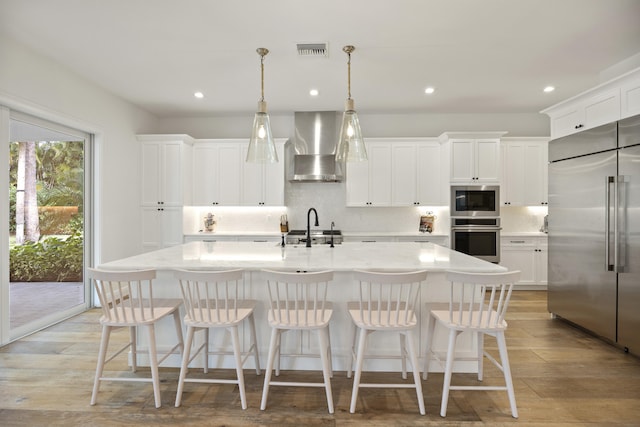 kitchen featuring an island with sink, sink, built in appliances, and wall chimney exhaust hood