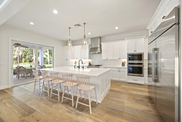 kitchen with decorative light fixtures, white cabinetry, high end refrigerator, a kitchen island with sink, and wall chimney exhaust hood