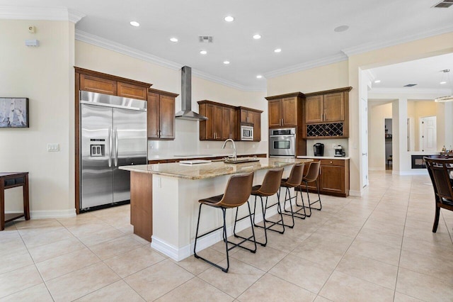 kitchen with a breakfast bar, a kitchen island with sink, built in appliances, light stone countertops, and wall chimney range hood