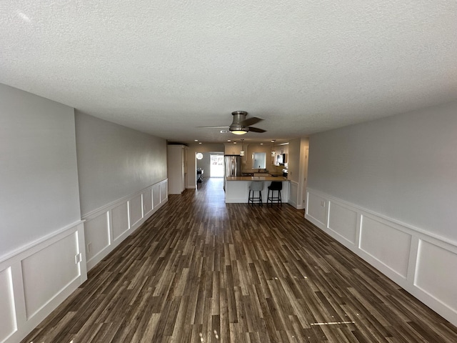 unfurnished living room featuring a decorative wall, a textured ceiling, dark wood-style flooring, a ceiling fan, and wainscoting