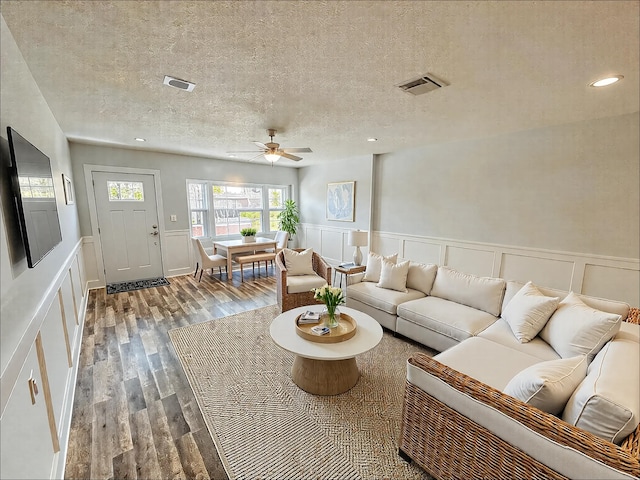 living room featuring a decorative wall, a ceiling fan, a textured ceiling, a wainscoted wall, and wood finished floors