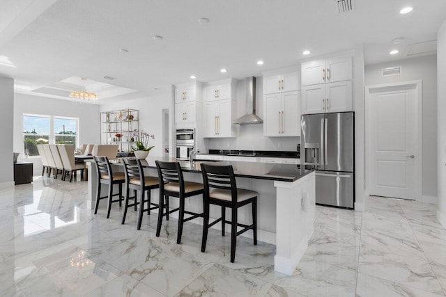 kitchen featuring wall chimney exhaust hood, a breakfast bar area, an island with sink, stainless steel appliances, and white cabinets