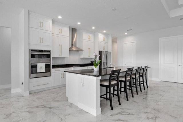 kitchen featuring appliances with stainless steel finishes, a breakfast bar area, white cabinets, a center island with sink, and wall chimney exhaust hood