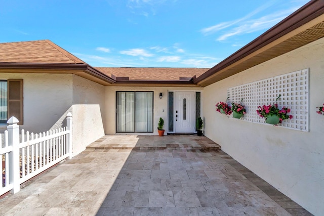 view of exterior entry featuring a patio area, a shingled roof, fence, and stucco siding