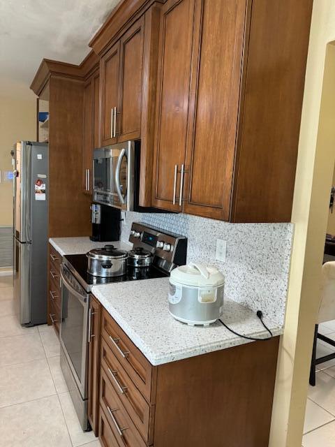 10298 Greentrail Dr N, Boynton Beach, FL 33436 - kitchen featuring appliances with stainless steel finishes, light tile patterned floors, decorative backsplash, light stone countertops, and brown cabinets