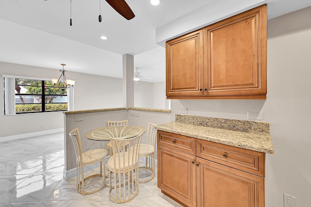 kitchen with light stone counters, marble finish floor, brown cabinetry, baseboards, and ceiling fan with notable chandelier