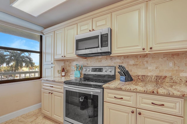 kitchen with cream cabinetry, light stone counters, tasteful backsplash, appliances with stainless steel finishes, and baseboards