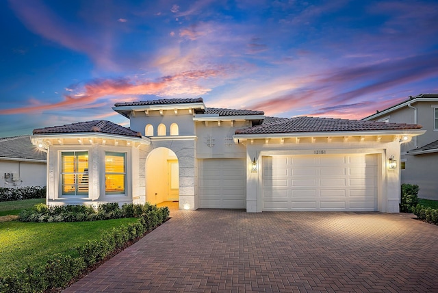 mediterranean / spanish house with a garage, a tiled roof, decorative driveway, a yard, and stucco siding