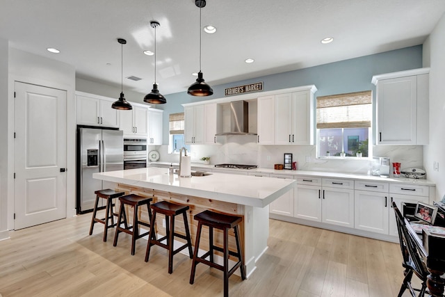 kitchen with white cabinetry, hanging light fixtures, a center island with sink, stainless steel appliances, and wall chimney range hood