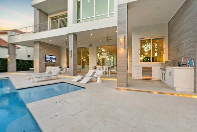 pool at dusk with exterior kitchen, area for grilling, ceiling fan, and a patio area