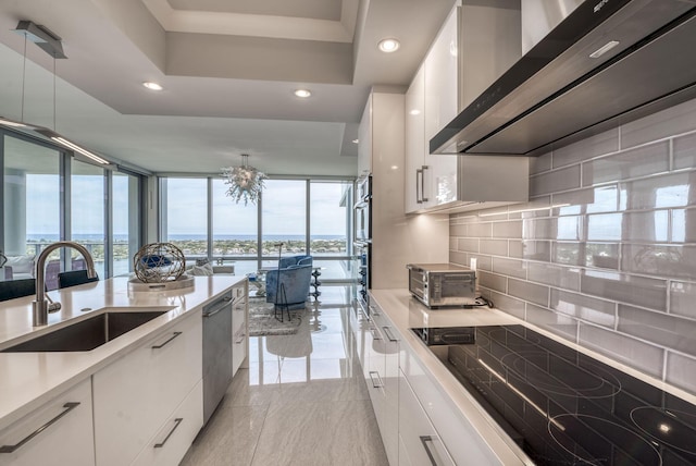 kitchen with decorative light fixtures, white cabinetry, sink, stainless steel dishwasher, and wall chimney range hood