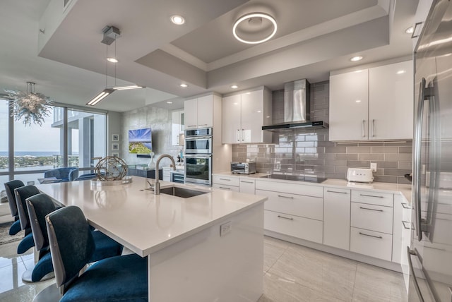 kitchen featuring a kitchen island with sink, sink, a raised ceiling, and wall chimney exhaust hood