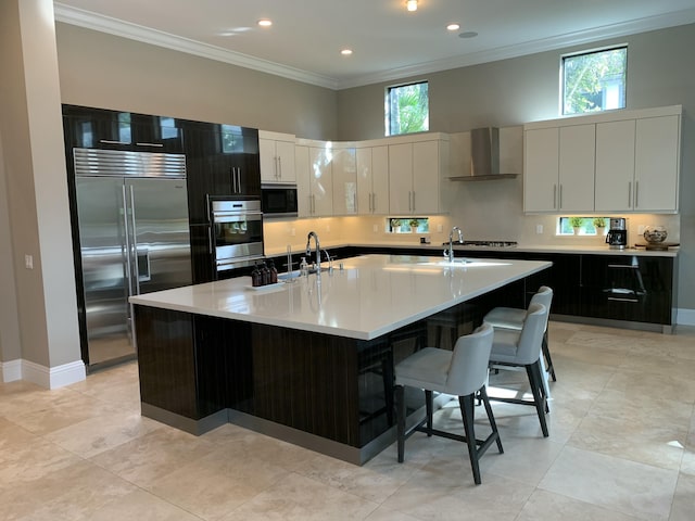 kitchen with a kitchen island with sink, built in appliances, white cabinets, and wall chimney exhaust hood