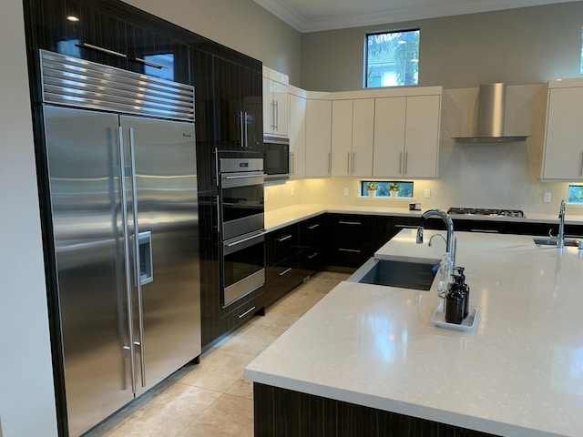 kitchen with sink, built in appliances, an island with sink, white cabinets, and wall chimney exhaust hood