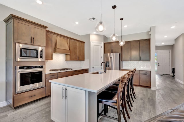 kitchen with sink, a kitchen island with sink, stainless steel appliances, decorative light fixtures, and custom exhaust hood