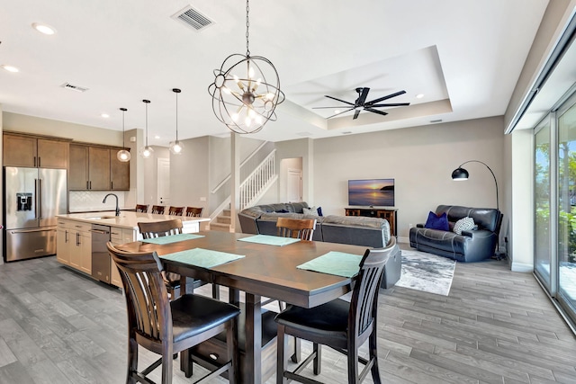 dining area featuring ceiling fan with notable chandelier, a raised ceiling, sink, and light wood-type flooring