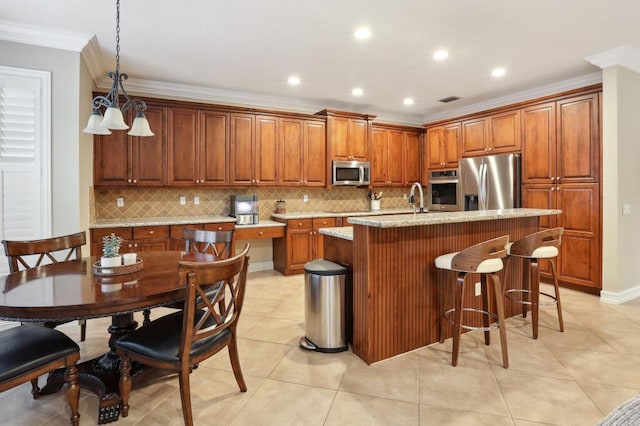 kitchen with decorative light fixtures, stainless steel appliances, crown molding, light stone countertops, and a center island with sink