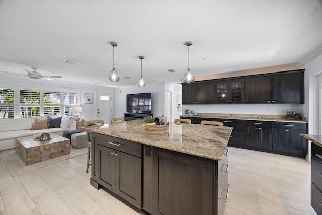 kitchen with decorative light fixtures, ornamental molding, a center island, light stone countertops, and dark brown cabinets