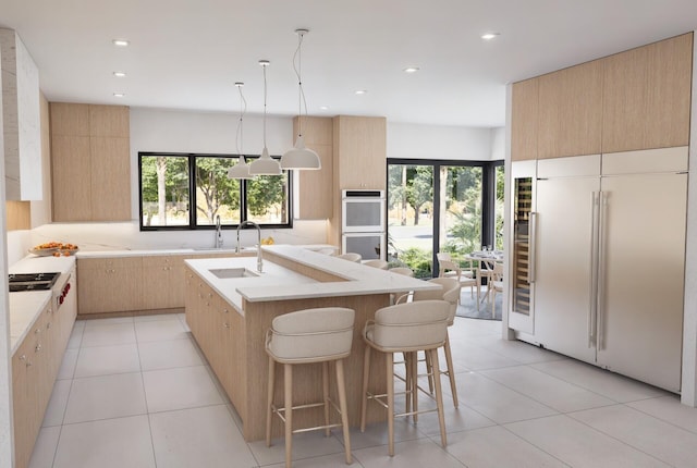 kitchen with hanging light fixtures, light brown cabinets, paneled built in refrigerator, double wall oven, and a kitchen island with sink