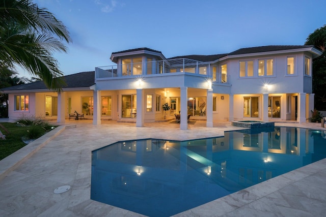 back house at dusk featuring a balcony, a pool with hot tub, and a patio