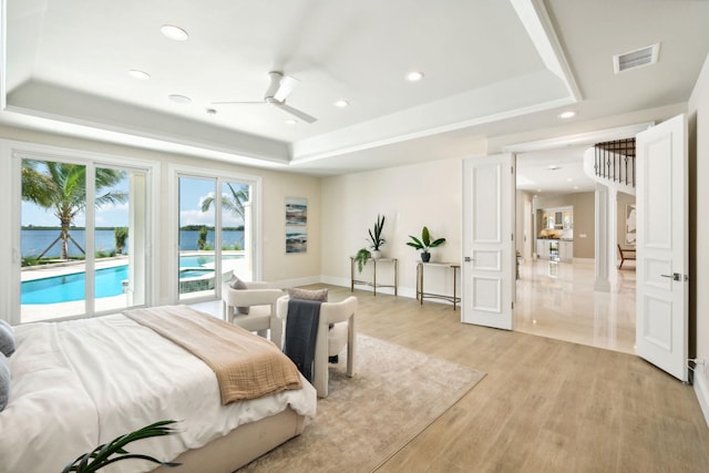 bedroom with a water view, light hardwood / wood-style flooring, a raised ceiling, ceiling fan, and access to exterior