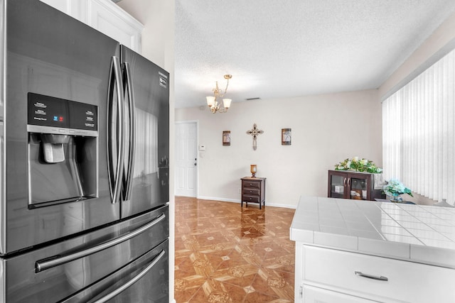 kitchen with tile counters, white cabinets, a textured ceiling, a notable chandelier, and stainless steel fridge