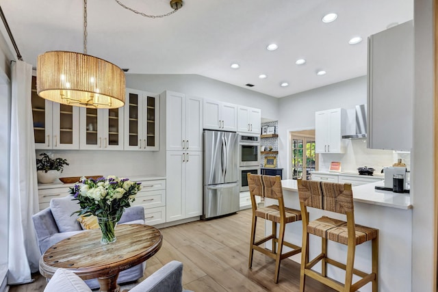 kitchen featuring wall chimney exhaust hood, vaulted ceiling, light hardwood / wood-style flooring, appliances with stainless steel finishes, and white cabinets