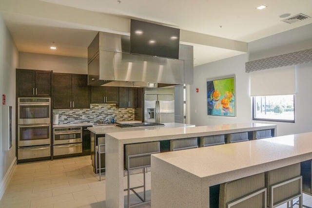 kitchen featuring appliances with stainless steel finishes, tasteful backsplash, a breakfast bar area, island exhaust hood, and a large island