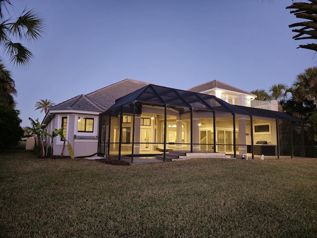 back house at dusk featuring a patio area, a lawn, and glass enclosure